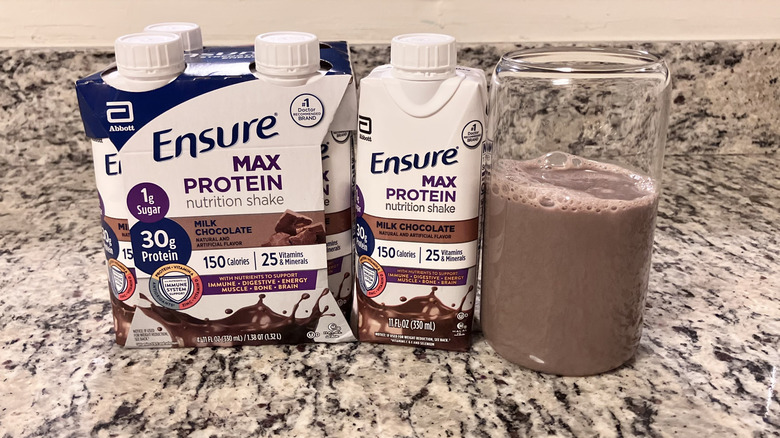A four-pack of Ensure Max Protein Nutrition Milk Chocolate Shake sits next to a bottle and clear glass containing the shake
