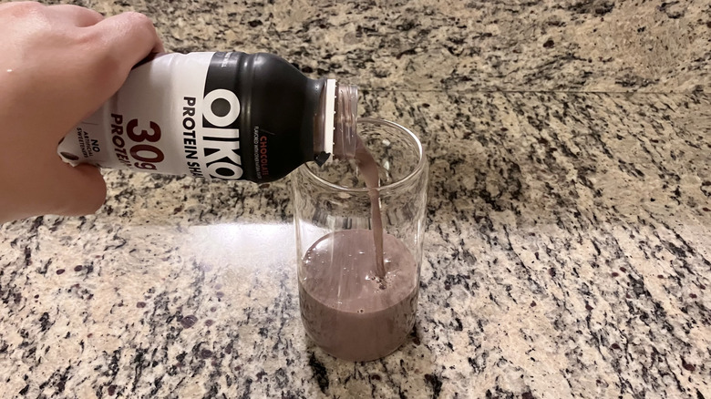 A person pours an OIKOS Chocolate Non-Caffeinated Casein Protein Shake into a clear glass on a counter
