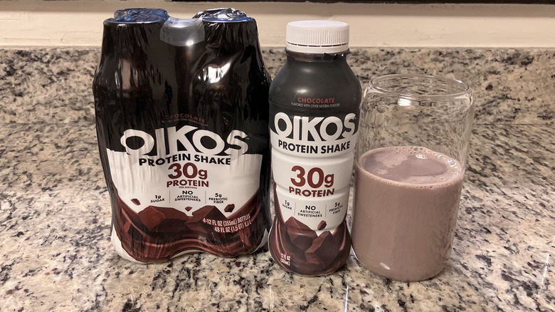 A four-pack of OIKOS Chocolate Non-Caffeinated Casein Protein Shakes sits next to a bottle and clear glass containing the shake