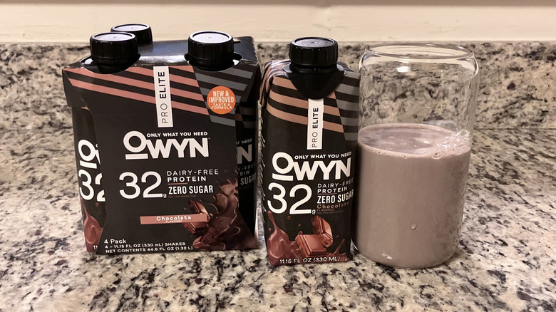 A four-pack of OWYN chocolate Pro Elite Vegan 32g Dairy-Free Protein Shakes sits next to a bottle and a clear glass containing the shake