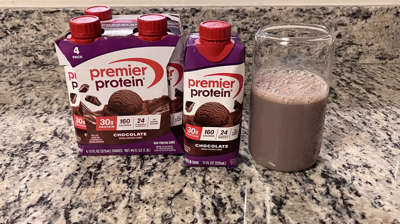 A four-pack of Premiere Protein Nutritional Chocolate Shakes sits next to a bottle and clear glass containing the shake