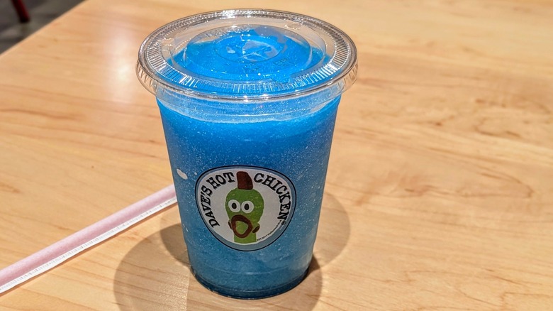 A blue slushie in a plastic cup next to a straw on a light-colored wood table
