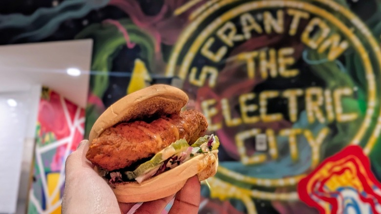A hand holding a chicken sandwich in front of a colorful mural at Dave's Hot Chicken