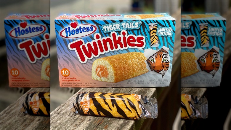 A box of Tiger Tails Twinkies featuring a golden sponge cake with orange filling. In front of it lies a Twinkie in transparent plastic wrap with a tiger stripe print on it