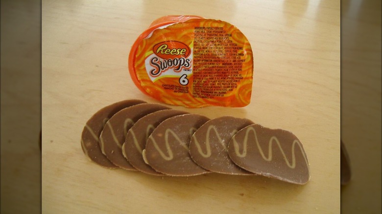 A line of brown chocolate Swoops (potato chip-shaped treats) with a lighter brown squiggly line down the center of each. They sit on a wooden table next to an orange, ovoid package that reads "Reese Swoops."