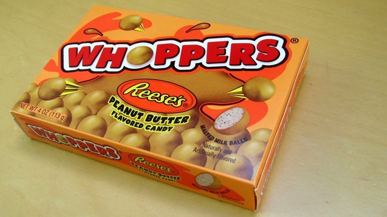 A box of Reese's Whoppers sitting on a wooden table. The box is orange, and depicts the brown-colored candies amid a simulated explosion of peanut butter.