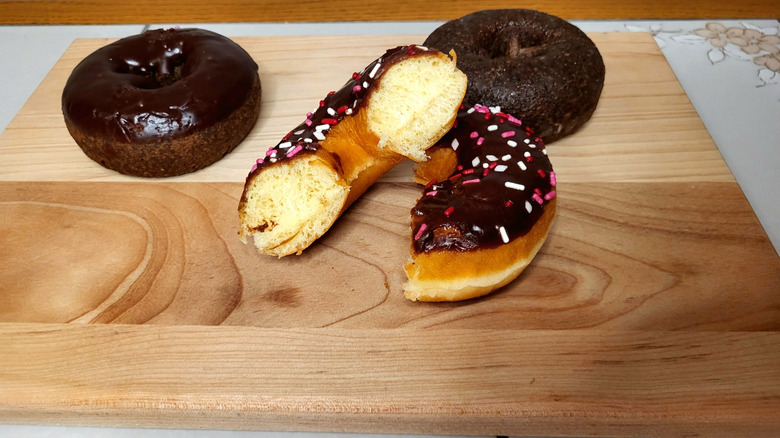A chocolate frosted donut cut in half, with one half lying on top of the other on a wooden cutting board. Two chocolate donuts sit behind it.