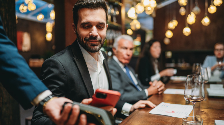 A man paying a bill with his red phone at a restaurant, people are in the background sat at the table