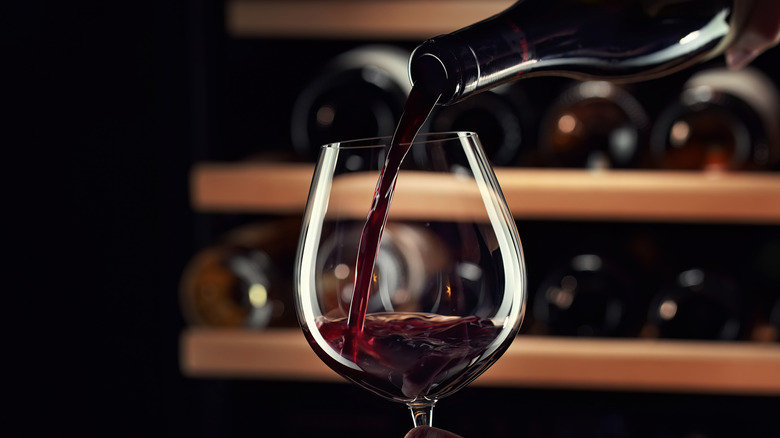 Close up of a person pouring red wine into a wine glass background of wine bottles of shelves