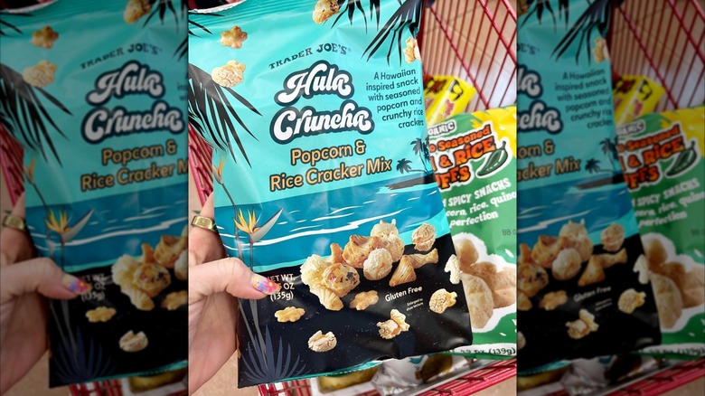 A hand holding a blue snack bag with a beach scene printed on it, accompanied by a mix of popcorn and rice crackers. On the bag are the words: "Trader Joe's Hula Cruncha Popcorn & Rice Cracker Mix."