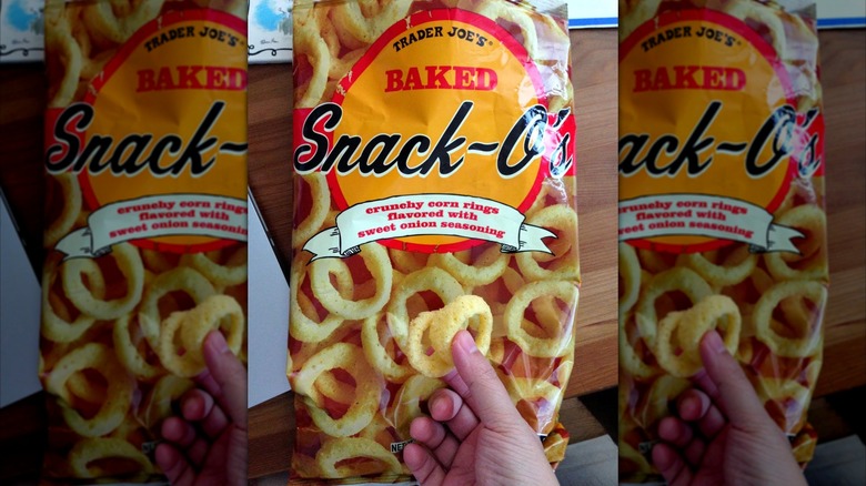 A hand holding a yellow onion ring snack in front of a bag of the same, with an image of more onion rings and the words "Trader Joe's Baked Snack-O's" printed in red and black text on a yellow backdrop.