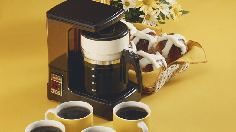 vintage 70s drop coffee maker on a "harvest gold" ground, with daisies and hot cross buns behind the pot and filled yellow coffee mugs in front