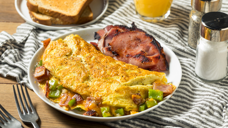Crispy Denver omelet stuffed with cheese, diced ham, and peppers with a side of charred ham on a white plate, sitting on a striped tablecloth with toast, orange juice, salt, and pepper