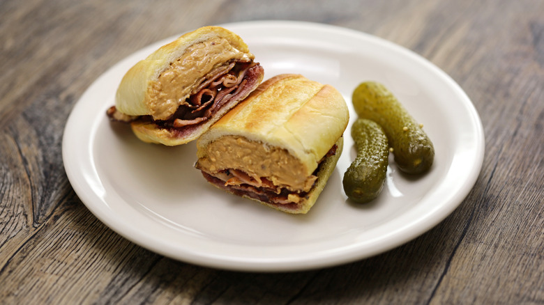 French bread sandwich containing peanut butter, jelly, and bacon served with pickles on a white plate on a wood table