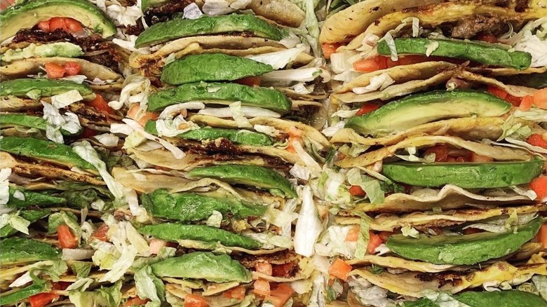 Tons of fried tacos topped with avocado jammed together, consisting of beef, tomatoes, lettuce, and cheese with a crispy shell