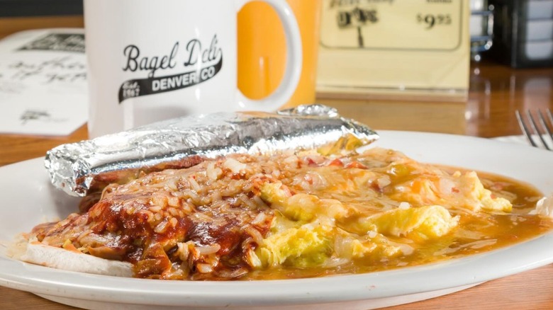 White plate with refried beans, lots of cheese, and red and green chile with a coffee cup reading "Bagel Deli" behind