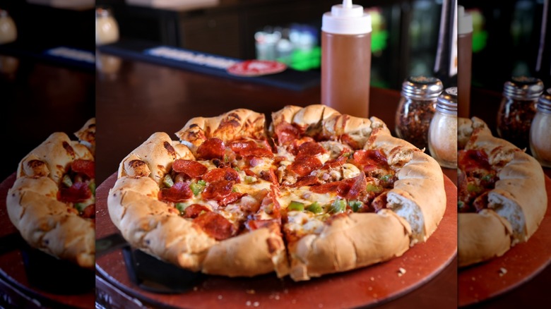 Hand-rolled, braided-crust Colorado pizza topped with pepperoni, cheese, and peppers on a wood serving platter with large bottle of honey and cheese and red pepper flake shakers behind it