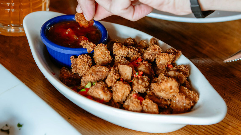 Hand holding a crispy Rocky Mountain oyster dipping into sweet chili sauce alongside a white, boat-like dish containing deep-fried Rocky Mountain oysters