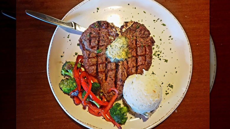 Beautiful grilled steak with char marks served with mashed potatoes, grilled peppers, and broccoli on a white plate against wood background