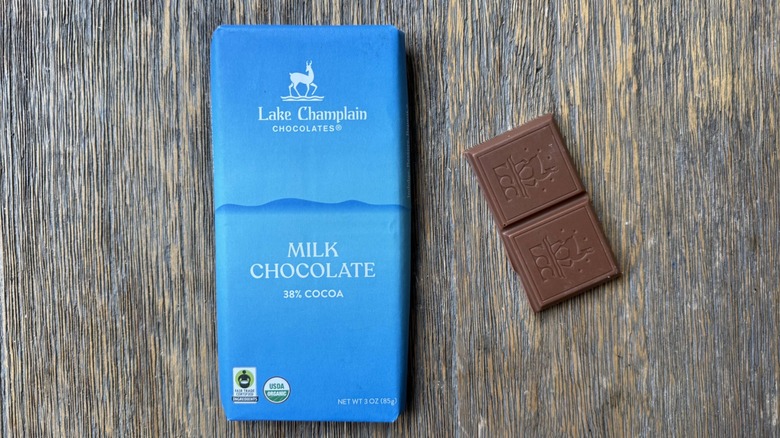 Wrapped Lake Champlain milk chocolate bar on table with rectangular piece beside it, consisting of two sections adorned with Lake Champlain Chocolates logo