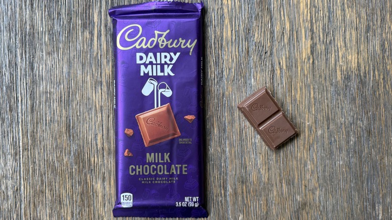 Wrapped Cadbury milk chocolate bar on table with small rectangular piece adjacent, consisting of two sections with Cadbury logo