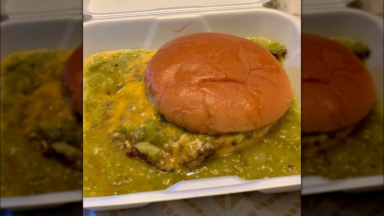 A burger in a white take-out container swimming in green chile sauce