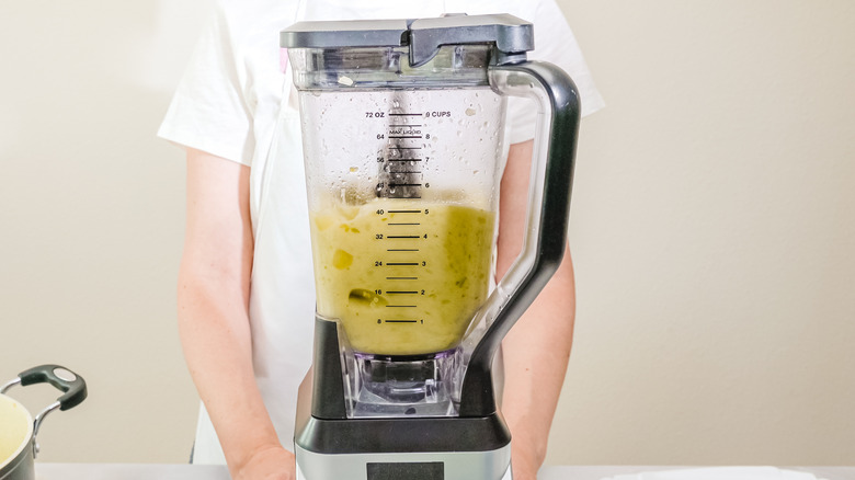 Celery soup in a jug blender with person standing behind it