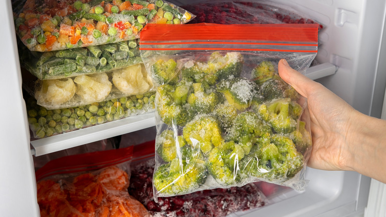 Hand removing bag of frozen broccoli from freezer with more frozen vegetables stacked in the back