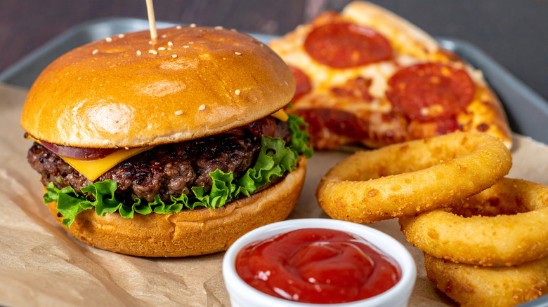 Cheeseburger, slice of pepperoni pizza, onion rings, and dish of ketchup on brown parchment paper