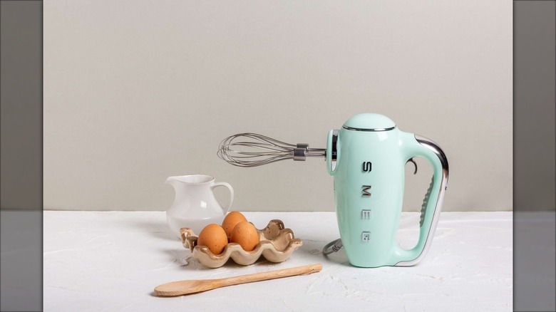 Light blue Smeg hand mixer sitting on table next to three eggs and milk pitcher