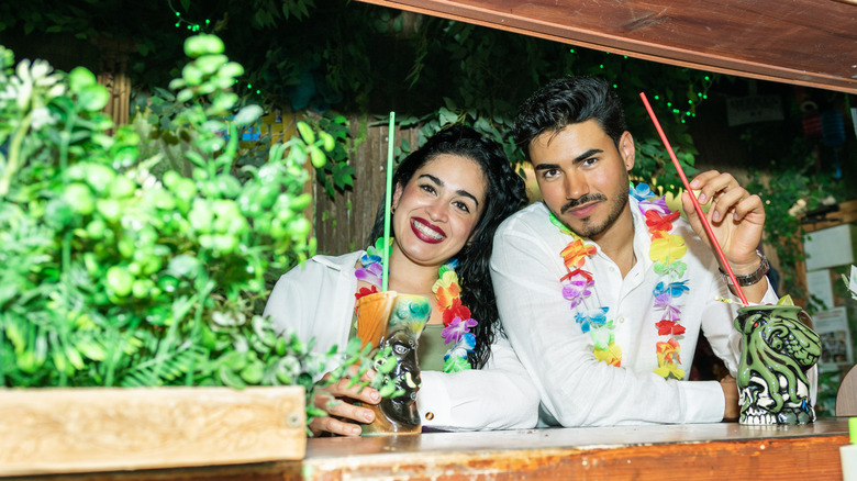 Two young adult bartenders smiling and posing for a photo, holding tiki cocktails in a tropical bar