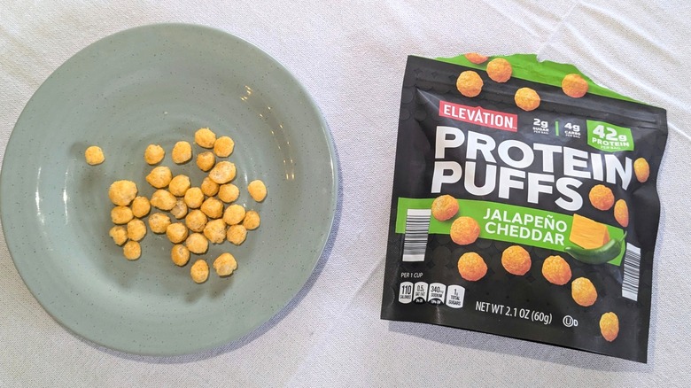 A bag of Elevation Jalapeño Cheddar Protein Puffs next to the puffs on a blue plate