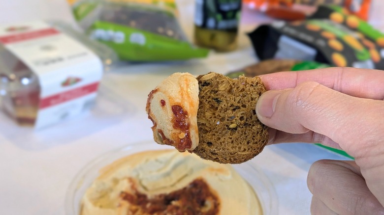 A close-up of a hand holding a cracker with spicy hummus and blurred Aldi products in the background