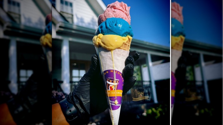 A gloved hand holds a slightly melting triple scoop cone, wrapped in white paper and shop branding.