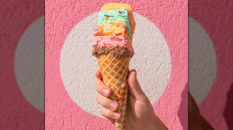 A hand holding a cone with a stack of multi-colored ice creams, in front of a pink wall.