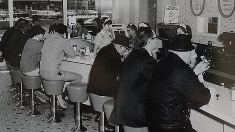 Black and white image of a 1970s Dunkin' Donuts with people sat at the counter