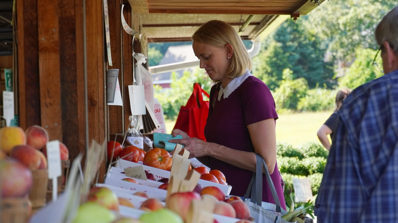 A woman takes out her wallet to pay at a farm stand with bags of apples
