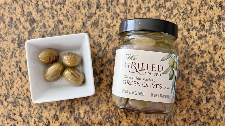 Jar of Grilled Pitted Green Olives with olives in a small dish
