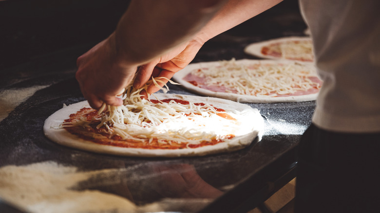 Man adding shredded mozzarella to pizza slathered with red sauce on work surface