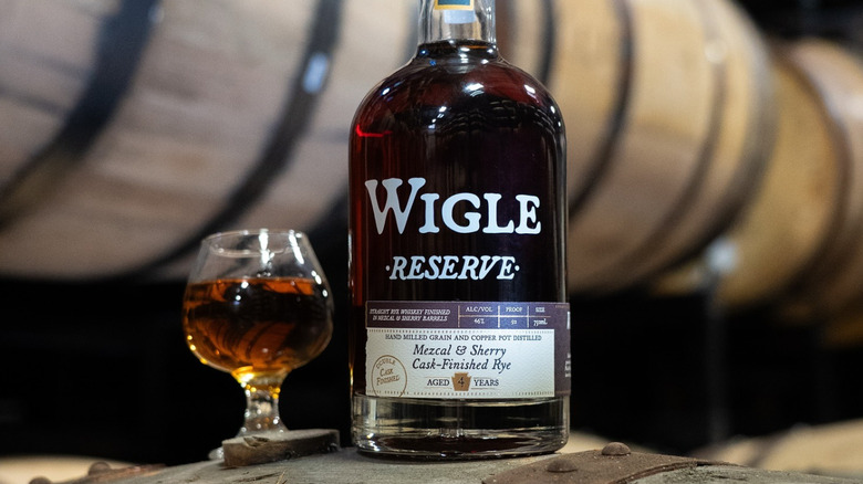 A close up of a bottle of Wigle Whiskey next to a filled glass with barrels in the background