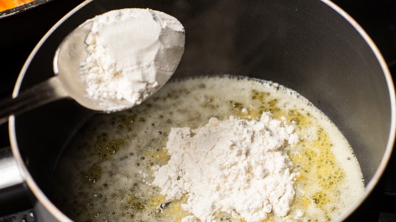 unseen hand adding a spoonful of flour to melted butter in a pan, making the roux as a base for a batch of bechamel sauce