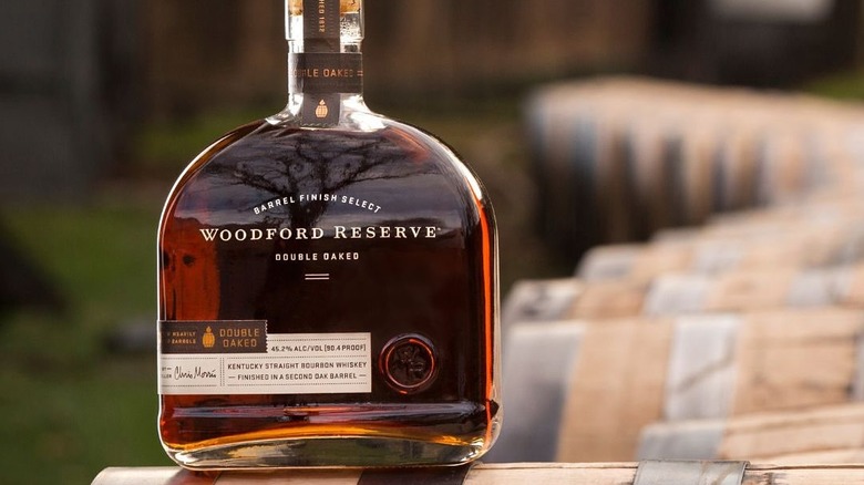 A close up of a bottle of Woodford Reserve Double Oaked sitting atop a whiskey barrel outdoors