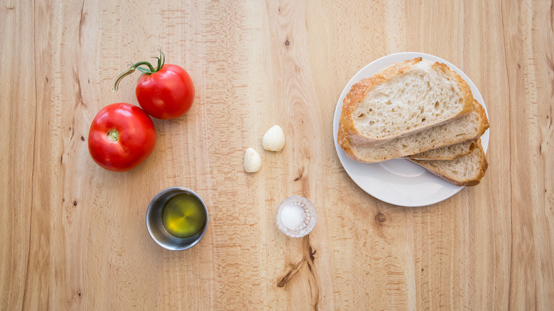 tomatoes, garlic cloves, bowl of salt, bowl of olive oil, and bread slices on plate laid out