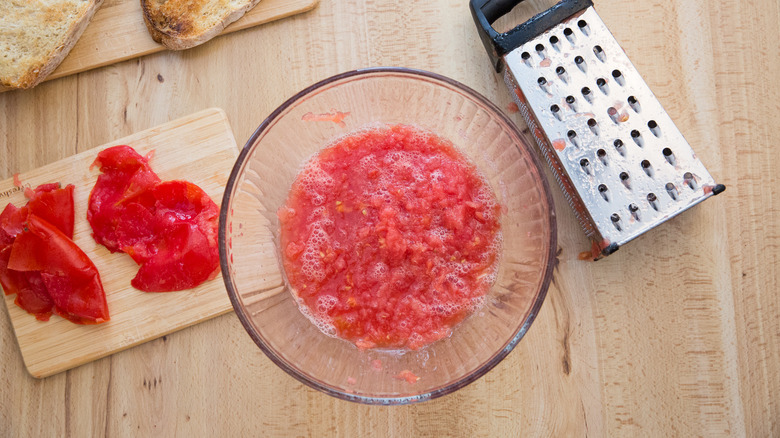 grated tomato in mixing bowl next to discarded skins on wooden board and grater