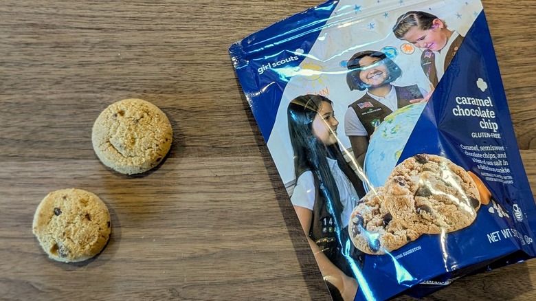 A bag of Caramel Chocolate Chip cookies next to two small cookies on a wood board