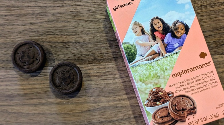 A box of Exploremores cookies next to two small, dark-brown cookies on a wood board
