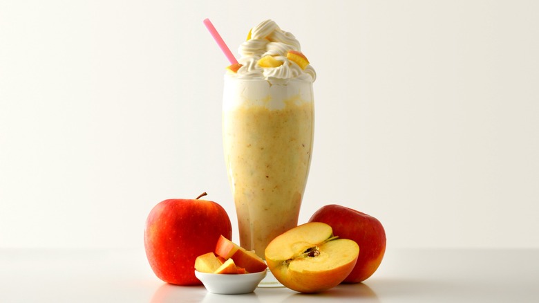 Apple milkshake in tall glass with whipped cream next to whole, halved, and chopped red apples