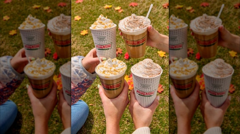Four hands holding various fall themed coffee drinks from Krispy Kreme over grass with autumn leaves