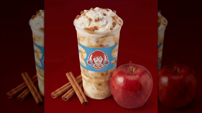 Wendy's Apple Crumble Frosty Fusion next to whole red apple and three cinnamon sticks on dark red background