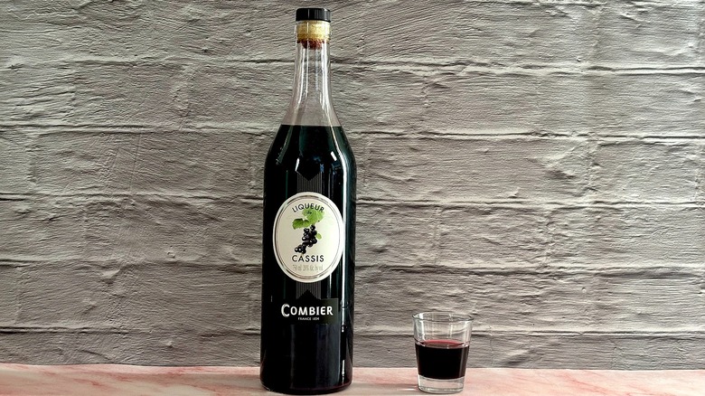 Bottle of Combier Liqueur de Cassis  next to shot glass of liqueur on grey brick background